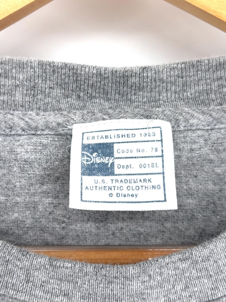 May include: A heather gray t-shirt featuring a white rectangular interior neck label with blue text that reads Established 1923, Disney, Code No. 78, Dept. 0018L, U.S. Trademark Authentic Clothing, and the Disney logo with a copyright symbol.