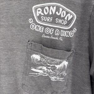 K&ouml;nnte beinhalten: Ein anthrazitfarbenes T-Shirt mit einer Brusttasche und einer wei&szlig;en Wellengrafik. &Uuml;ber der Tasche steht in wei&szlig;em Text RON JON SURF SHOP, ONE OF A KIND und Cocoa Beach, Fla. Der Stoff des Kleidungsst&uuml;cks weist eine melierte Textur auf.