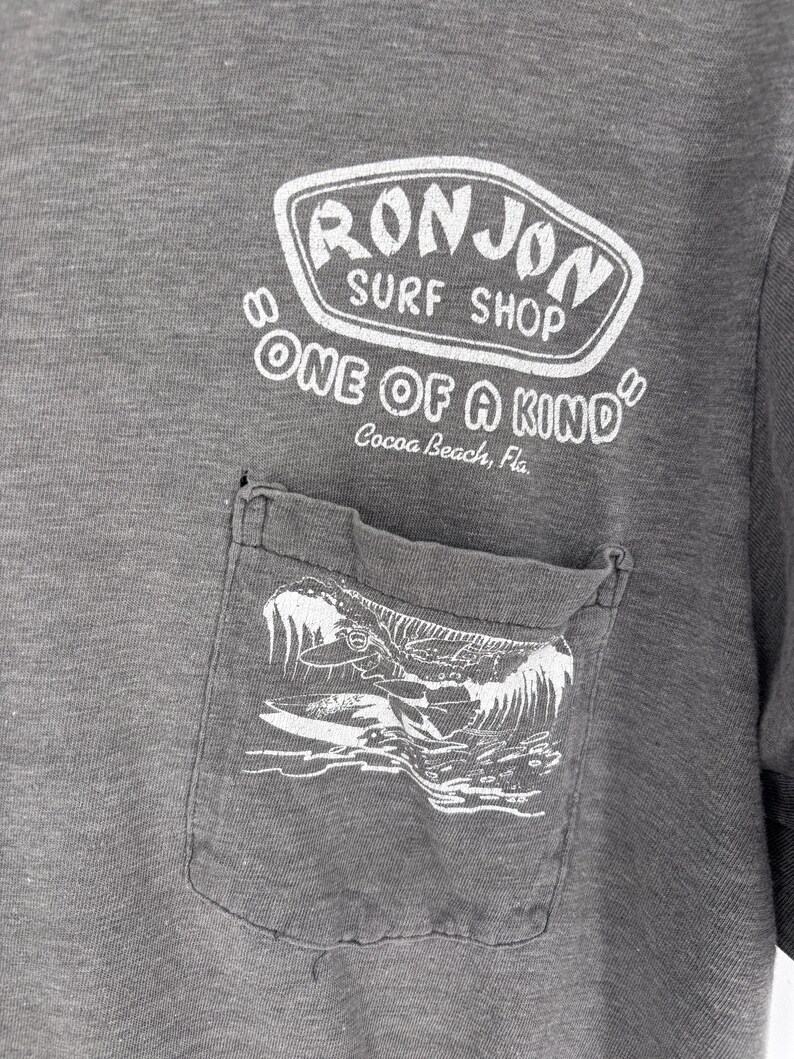 K&ouml;nnte beinhalten: Ein anthrazitfarbenes T-Shirt mit einer Brusttasche und einer wei&szlig;en Wellengrafik. &Uuml;ber der Tasche steht in wei&szlig;em Text RON JON SURF SHOP, ONE OF A KIND und Cocoa Beach, Fla. Der Stoff des Kleidungsst&uuml;cks weist eine melierte Textur auf.
