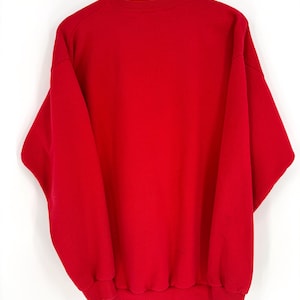 K&ouml;nnte beinhalten: Ein einfarbiges rotes Sweatshirt, das an einem Holzb&uuml;gel h&auml;ngt. Das Sweatshirt hat einen Rundhalsausschnitt und lange &Auml;rmel mit gerippten B&uuml;ndchen und Saum. Das Kleidungsst&uuml;ck besteht aus einem weichen, dicken Material.