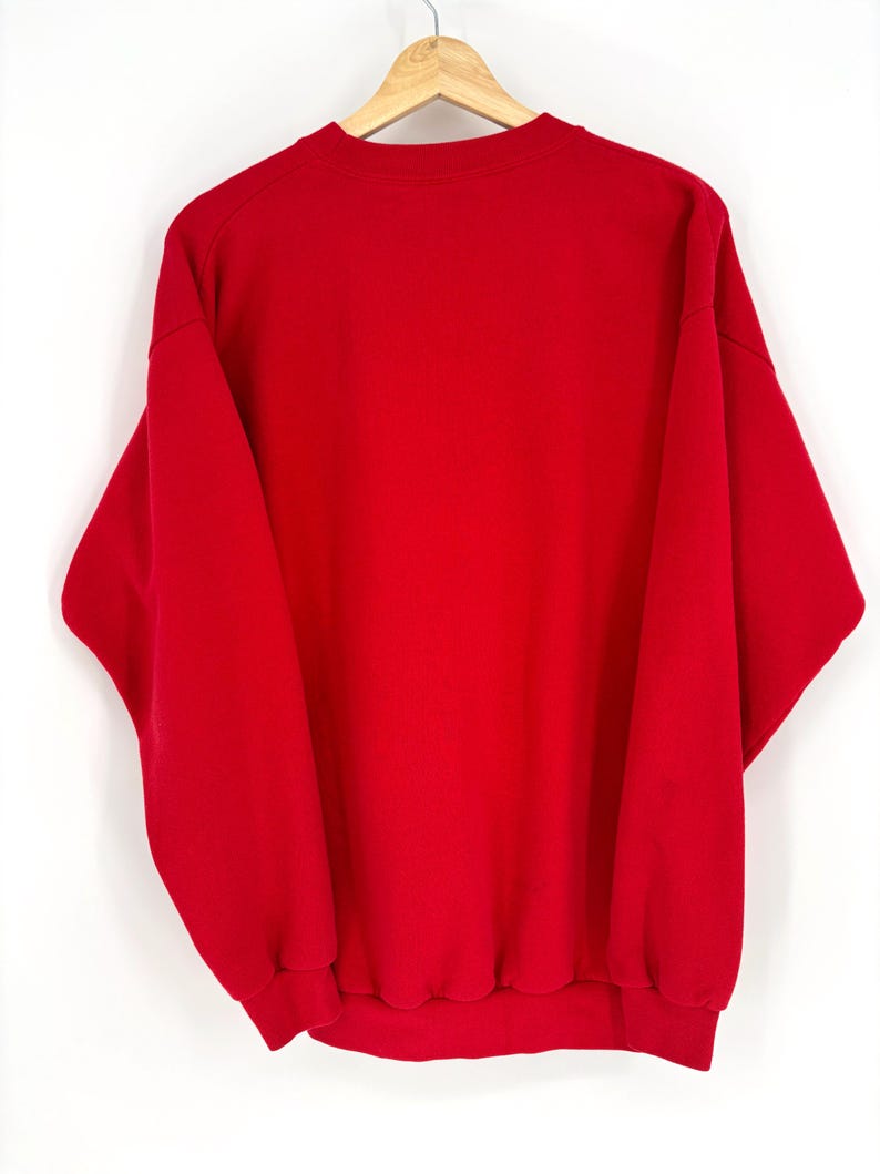 K&ouml;nnte beinhalten: Ein einfarbiges rotes Sweatshirt, das an einem Holzb&uuml;gel h&auml;ngt. Das Sweatshirt hat einen Rundhalsausschnitt und lange &Auml;rmel mit gerippten B&uuml;ndchen und Saum. Das Kleidungsst&uuml;ck besteht aus einem weichen, dicken Material.