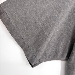 K&ouml;nnte beinhalten: Ein kurz&auml;rmeliges T-Shirt aus meliertem anthrazitfarbenem Stoff. Das Kleidungsst&uuml;ck weist einen sichtbaren Doppeln&auml;htsaum entlang der &Auml;rmelkante auf, der die Textur des Strickmaterials und die Konstruktion der &Auml;rmelnaht deutlich hervorhebt.