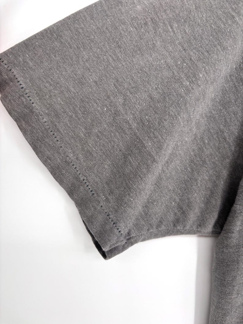 K&ouml;nnte beinhalten: Ein kurz&auml;rmeliges T-Shirt aus meliertem anthrazitfarbenem Stoff. Das Kleidungsst&uuml;ck weist einen sichtbaren Doppeln&auml;htsaum entlang der &Auml;rmelkante auf, der die Textur des Strickmaterials und die Konstruktion der &Auml;rmelnaht deutlich hervorhebt.