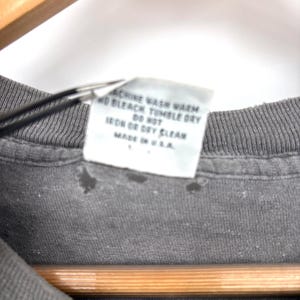 K&ouml;nnte beinhalten: Ein dunkelgraues T-Shirt mit einem wei&szlig;en Pflegeetikett an der Innenseite, das den Text enth&auml;lt: MACHINE WASH WARM, NO BLEACH, TUMBLE DRY, DO NOT IRON OR DRY CLEAN, MADE IN U.S.A. Der Stoff zeigt leichte Abnutzungserscheinungen am Kragen.