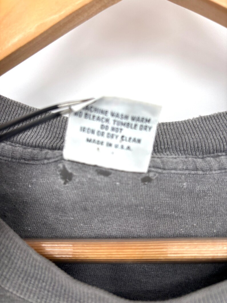 K&ouml;nnte beinhalten: Ein dunkelgraues T-Shirt mit einem wei&szlig;en Pflegeetikett an der Innenseite, das den Text enth&auml;lt: MACHINE WASH WARM, NO BLEACH, TUMBLE DRY, DO NOT IRON OR DRY CLEAN, MADE IN U.S.A. Der Stoff zeigt leichte Abnutzungserscheinungen am Kragen.