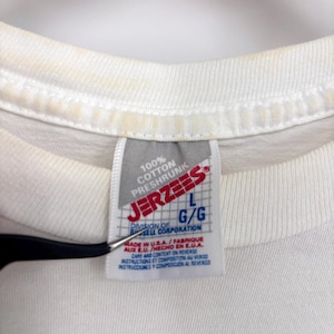 May include: A white t-shirt featuring a Jerzees brand tag at the neckline. The tag displays the text 100% Cotton Preshrunk, Jerzees, L G/G, Division of Russell Corporation, Made in U.S.A., and Care and Content on Reverse in English, French, and Spanish.