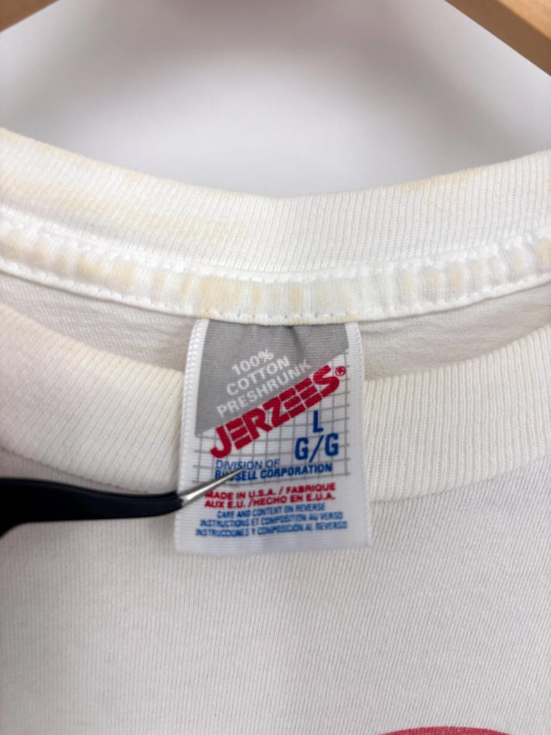 May include: A white t-shirt featuring a Jerzees brand tag at the neckline. The tag displays the text 100% Cotton Preshrunk, Jerzees, L G/G, Division of Russell Corporation, Made in U.S.A., and Care and Content on Reverse in English, French, and Spanish.