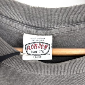 K&ouml;nnte beinhalten: Ein graues T-Shirt mit einem wei&szlig;en Innenetikett am Kragen, das den Text 100% COTTON PRESHRUNK, RONJON SURF T'S und LARGE enth&auml;lt. Das Etikett ist rechteckig mit einem rot-schwarzen Rand, und das T-Shirt hat einen gerippten Rundhalsausschnitt.