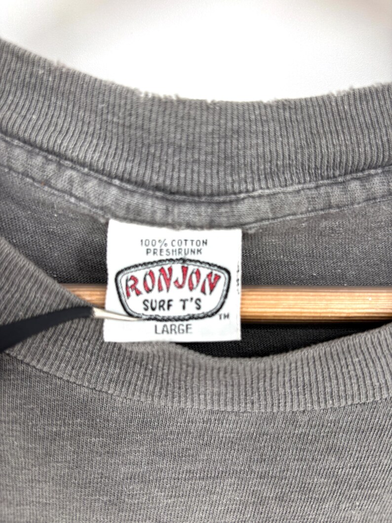 K&ouml;nnte beinhalten: Ein graues T-Shirt mit einem wei&szlig;en Innenetikett am Kragen, das den Text 100% COTTON PRESHRUNK, RONJON SURF T'S und LARGE enth&auml;lt. Das Etikett ist rechteckig mit einem rot-schwarzen Rand, und das T-Shirt hat einen gerippten Rundhalsausschnitt.