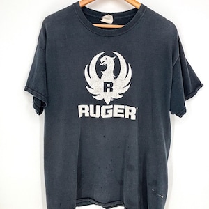 May include: A faded black short-sleeve t-shirt featuring a white graphic of the Ruger eagle logo and the text RUGER printed in white across the chest. The shirt has a crew neckline and shows signs of wear with small holes and light discoloration.