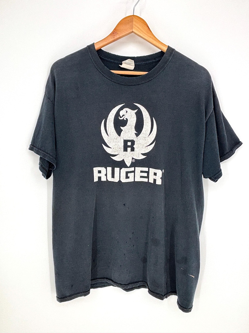 May include: A faded black short-sleeve t-shirt featuring a white graphic of the Ruger eagle logo and the text RUGER printed in white across the chest. The shirt has a crew neckline and shows signs of wear with small holes and light discoloration.
