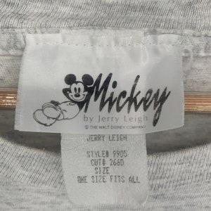 Puede incluir: Etiqueta blanca con un personaje de dibujos animados de Mickey Mouse negro y el texto "Mickey by Jerry Leigh", "The Walt Disney Company", "Jerry Leigh", "Style# 9905", "Cut# 2660", "Size", "One Size Fits All".