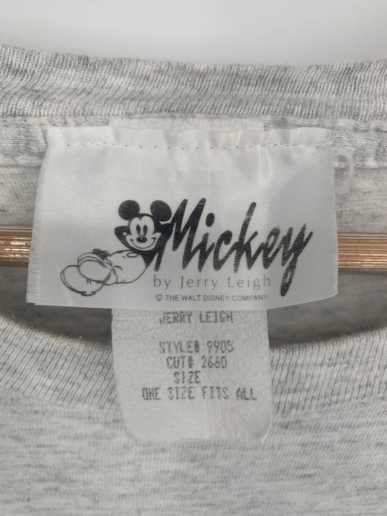 Puede incluir: Etiqueta blanca con un personaje de dibujos animados de Mickey Mouse negro y el texto "Mickey by Jerry Leigh", "The Walt Disney Company", "Jerry Leigh", "Style# 9905", "Cut# 2660", "Size", "One Size Fits All".