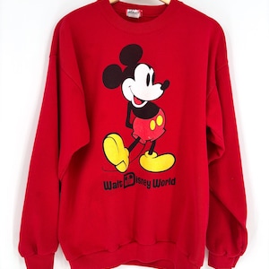 K&ouml;nnte beinhalten: Ein rotes Sweatshirt mit einer Mickey Mouse-Grafik. Die Zeichentrickfigur ist mit schwarzen Ohren, einem wei&szlig;en Gesicht, roten Shorts und gelben Schuhen dargestellt. Die Worte "Walt Disney World" sind darunter aufgedruckt.