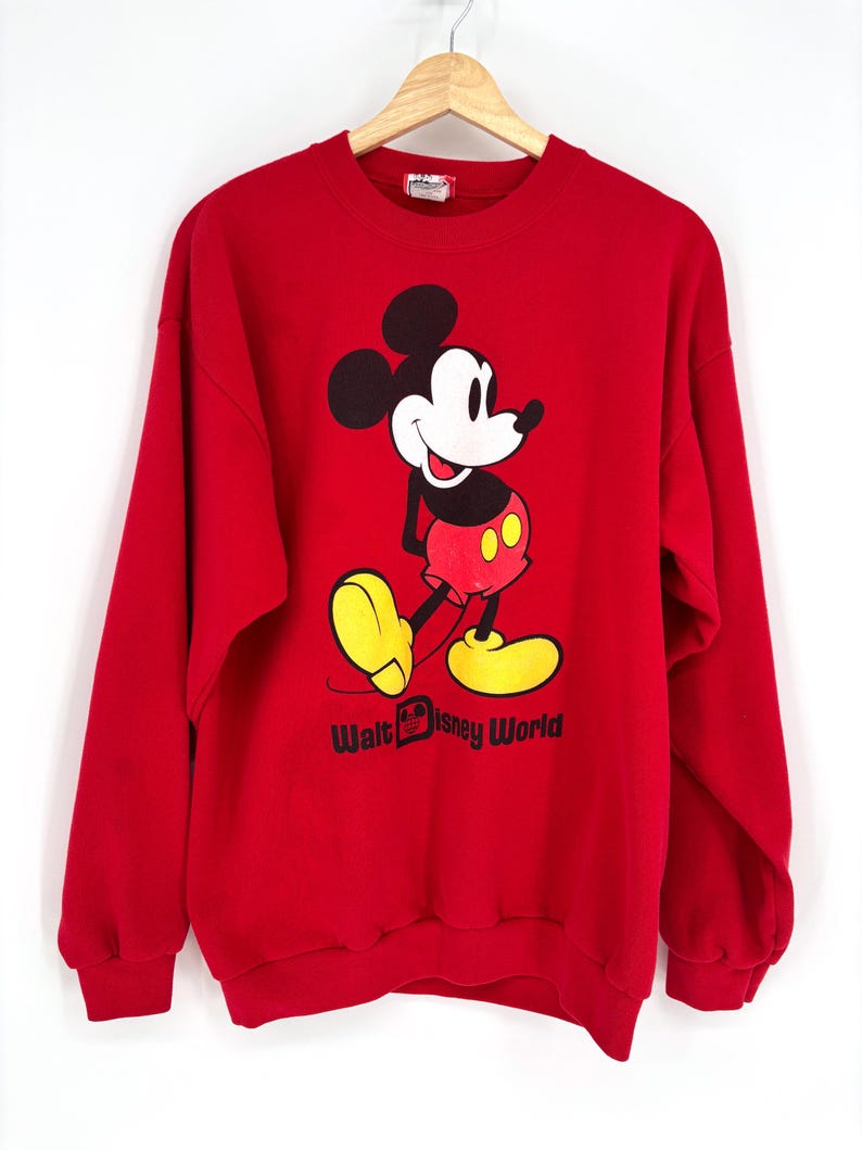 K&ouml;nnte beinhalten: Ein rotes Sweatshirt mit einer Mickey Mouse-Grafik. Die Zeichentrickfigur ist mit schwarzen Ohren, einem wei&szlig;en Gesicht, roten Shorts und gelben Schuhen dargestellt. Die Worte "Walt Disney World" sind darunter aufgedruckt.