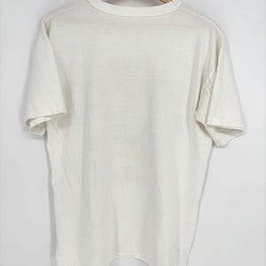 May include: A plain off-white short-sleeve t-shirt with a crew neckline. The garment is displayed hanging on a wooden hanger, featuring a simple, solid-colored design with short sleeves and a standard hemline, presented against a neutral background.