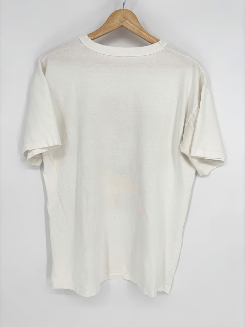 May include: A plain off-white short-sleeve t-shirt with a crew neckline. The garment is displayed hanging on a wooden hanger, featuring a simple, solid-colored design with short sleeves and a standard hemline, presented against a neutral background.