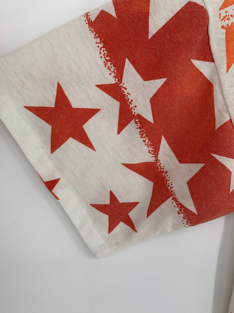 Op de afbeelding: Close-up van een witte stof met een patroon van oranje sterren. De stof heeft een licht getextureerd oppervlak.