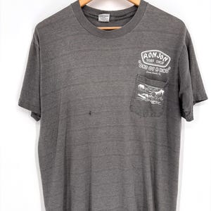 K&ouml;nnte beinhalten: Ein anthrazitfarbenes Kurzarm-T-Shirt mit einer Brusttasche, die eine wei&szlig;e Grafik eines Surfers auf einer Welle zeigt. &Uuml;ber der Tasche steht der Text RON JON SURF SHOP ONE OF A KIND Cocoa Beach, FL. Das Shirt hat einen Rundhalsausschnitt und ein verwaschenes Finish.