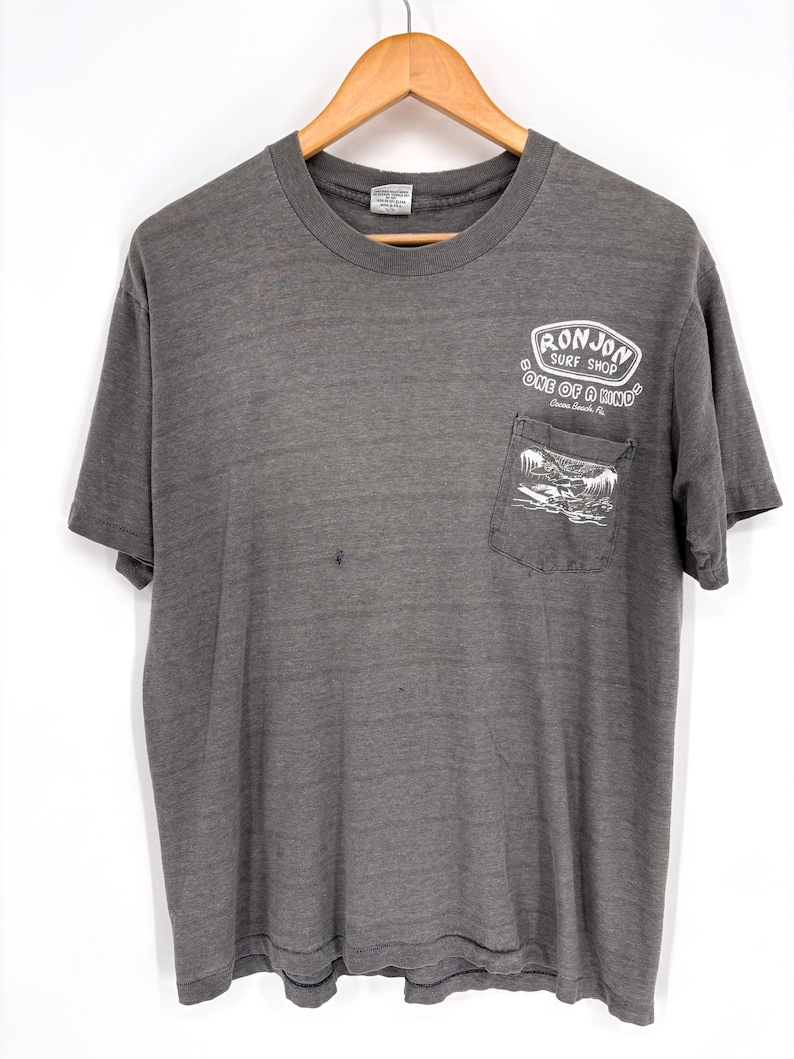 K&ouml;nnte beinhalten: Ein anthrazitfarbenes Kurzarm-T-Shirt mit einer Brusttasche, die eine wei&szlig;e Grafik eines Surfers auf einer Welle zeigt. &Uuml;ber der Tasche steht der Text RON JON SURF SHOP ONE OF A KIND Cocoa Beach, FL. Das Shirt hat einen Rundhalsausschnitt und ein verwaschenes Finish.