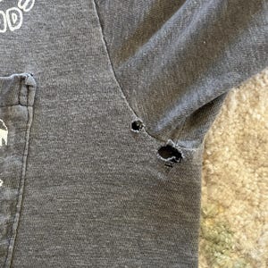 K&ouml;nnte beinhalten: Ein anthrazitfarbenes T-Shirt mit Brusttasche und wei&szlig;em Textaufdruck. Das Kleidungsst&uuml;ck weist zwei sichtbare L&ouml;cher entlang der Achselnaht auf, die Abnutzungserscheinungen und Ausfransungen des Stoffes am &Auml;rmelansatz und der Seitennaht zeigen.