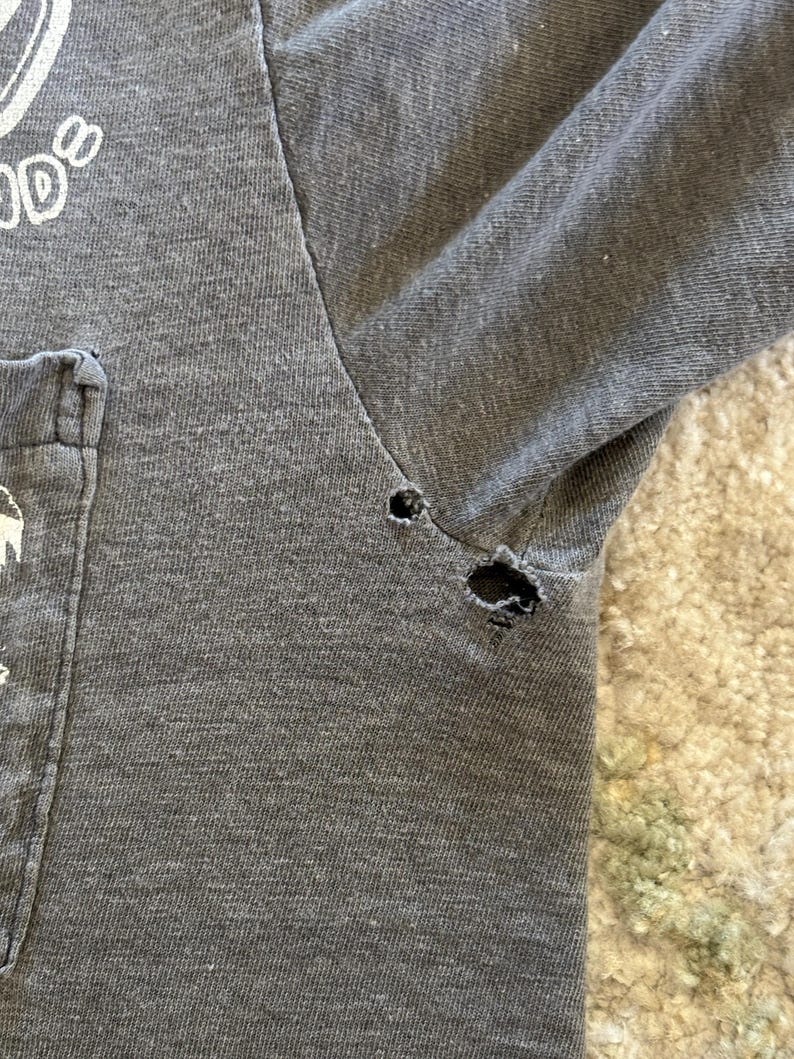 K&ouml;nnte beinhalten: Ein anthrazitfarbenes T-Shirt mit Brusttasche und wei&szlig;em Textaufdruck. Das Kleidungsst&uuml;ck weist zwei sichtbare L&ouml;cher entlang der Achselnaht auf, die Abnutzungserscheinungen und Ausfransungen des Stoffes am &Auml;rmelansatz und der Seitennaht zeigen.