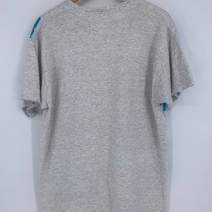 Puede incluir: Una camiseta gris jaspeada con un estampado gr&aacute;fico azul y blanco en la parte delantera y trasera. El estampado gr&aacute;fico presenta un dise&ntilde;o estilizado de una persona sosteniendo un micr&oacute;fono.