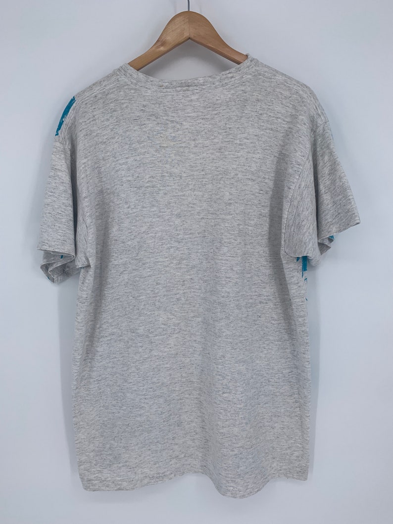 Puede incluir: Una camiseta gris jaspeada con un estampado gr&aacute;fico azul y blanco en la parte delantera y trasera. El estampado gr&aacute;fico presenta un dise&ntilde;o estilizado de una persona sosteniendo un micr&oacute;fono.