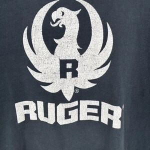 May include: A dark charcoal gray t-shirt featuring a large, distressed white graphic of the Ruger eagle logo centered above the word RUGER in bold, block lettering. The fabric shows signs of wear with small holes visible near the bottom hem.