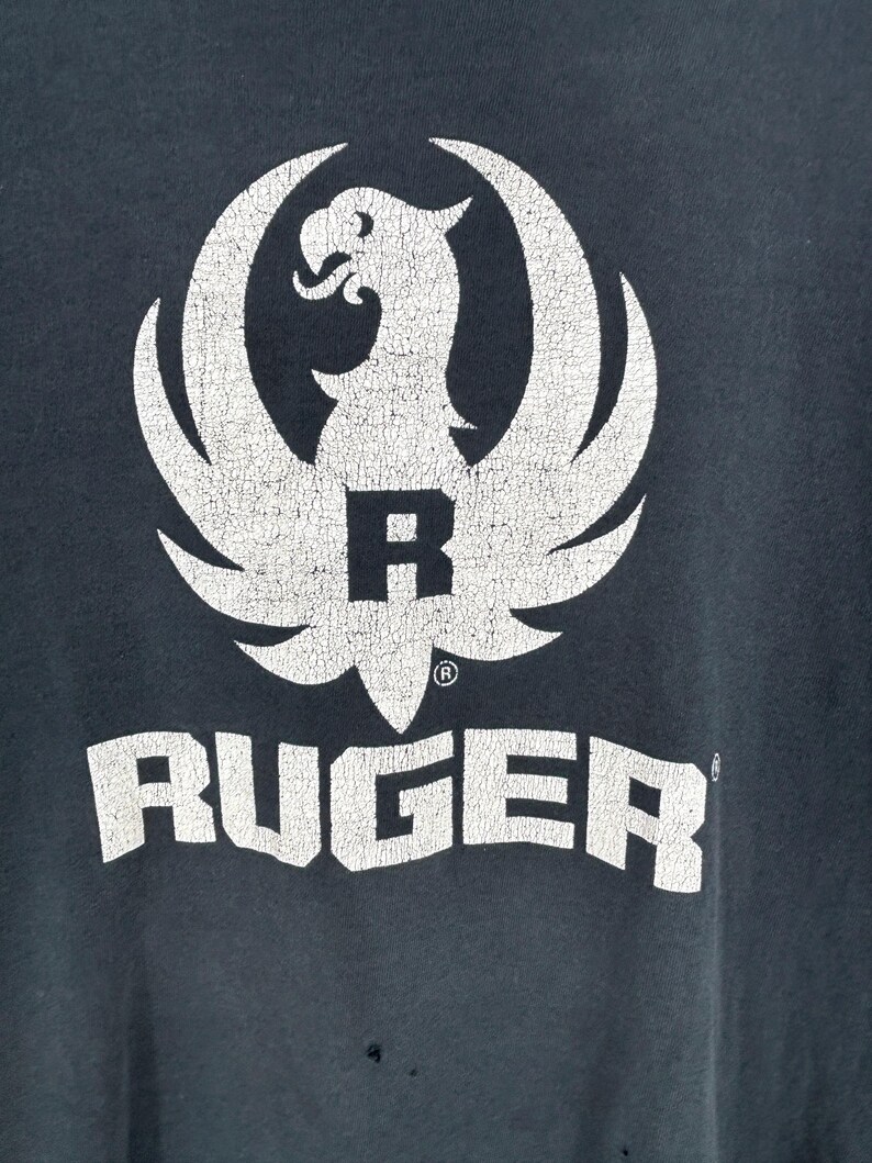 May include: A dark charcoal gray t-shirt featuring a large, distressed white graphic of the Ruger eagle logo centered above the word RUGER in bold, block lettering. The fabric shows signs of wear with small holes visible near the bottom hem.