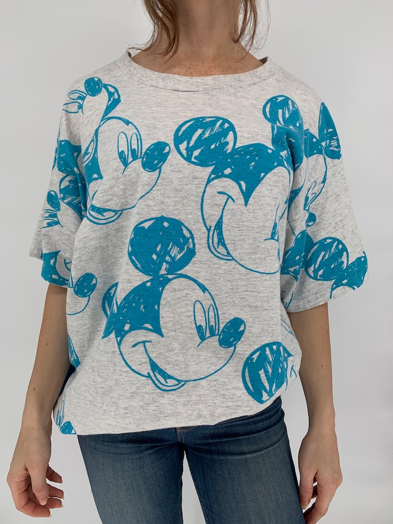 Puede incluir: Una camiseta gris con un estampado azul de Mickey Mouse. El estampado es un estilo de dibujo del rostro de Mickey Mouse.