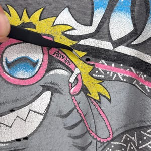 K&ouml;nnte beinhalten: Ein graues T-Shirt mit einem Grafikdruck einer Zeichentrickfigur mit gelben Haaren, rosa Brille und einem breiten L&auml;cheln. Das Design enth&auml;lt den Text "RIWJIN" auf dem rosa Brillenrahmen, mit sichtbaren Rissen in der Druckfarbe.