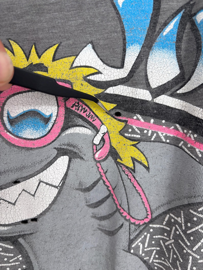 K&ouml;nnte beinhalten: Ein graues T-Shirt mit einem Grafikdruck einer Zeichentrickfigur mit gelben Haaren, rosa Brille und einem breiten L&auml;cheln. Das Design enth&auml;lt den Text "RIWJIN" auf dem rosa Brillenrahmen, mit sichtbaren Rissen in der Druckfarbe.