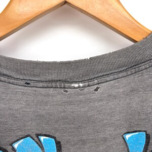K&ouml;nnte beinhalten: Ein anthrazitfarbenes T-Shirt mit einem Grafikdruck in leuchtendem Blau und Gelb. Der gerippte Rundhalsausschnitt weist Gebrauchsspuren mit kleinen L&ouml;chern entlang der Naht auf. Der Stoff hat eine melierte Textur und die Grafik enth&auml;lt stilisierte Schrift.
