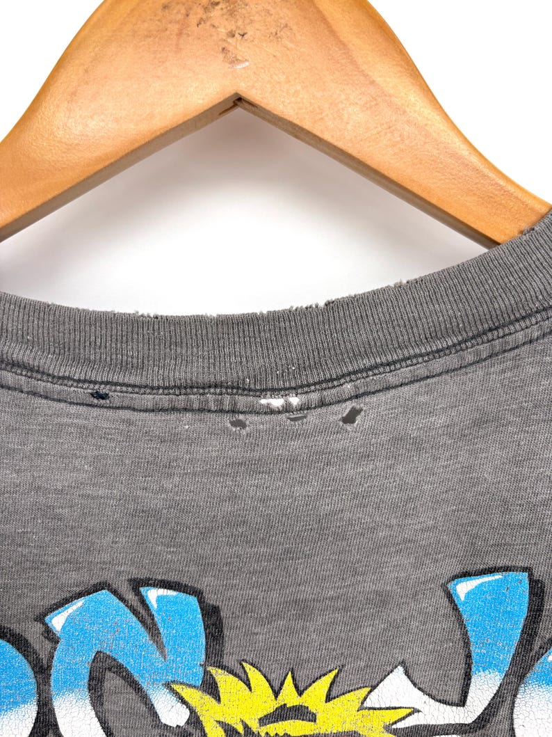 K&ouml;nnte beinhalten: Ein anthrazitfarbenes T-Shirt mit einem Grafikdruck in leuchtendem Blau und Gelb. Der gerippte Rundhalsausschnitt weist Gebrauchsspuren mit kleinen L&ouml;chern entlang der Naht auf. Der Stoff hat eine melierte Textur und die Grafik enth&auml;lt stilisierte Schrift.