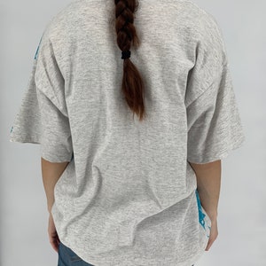 Puede incluir: Una camiseta gris con un estampado gr&aacute;fico azul en la espalda. La camiseta tiene cuello redondo y mangas cortas.