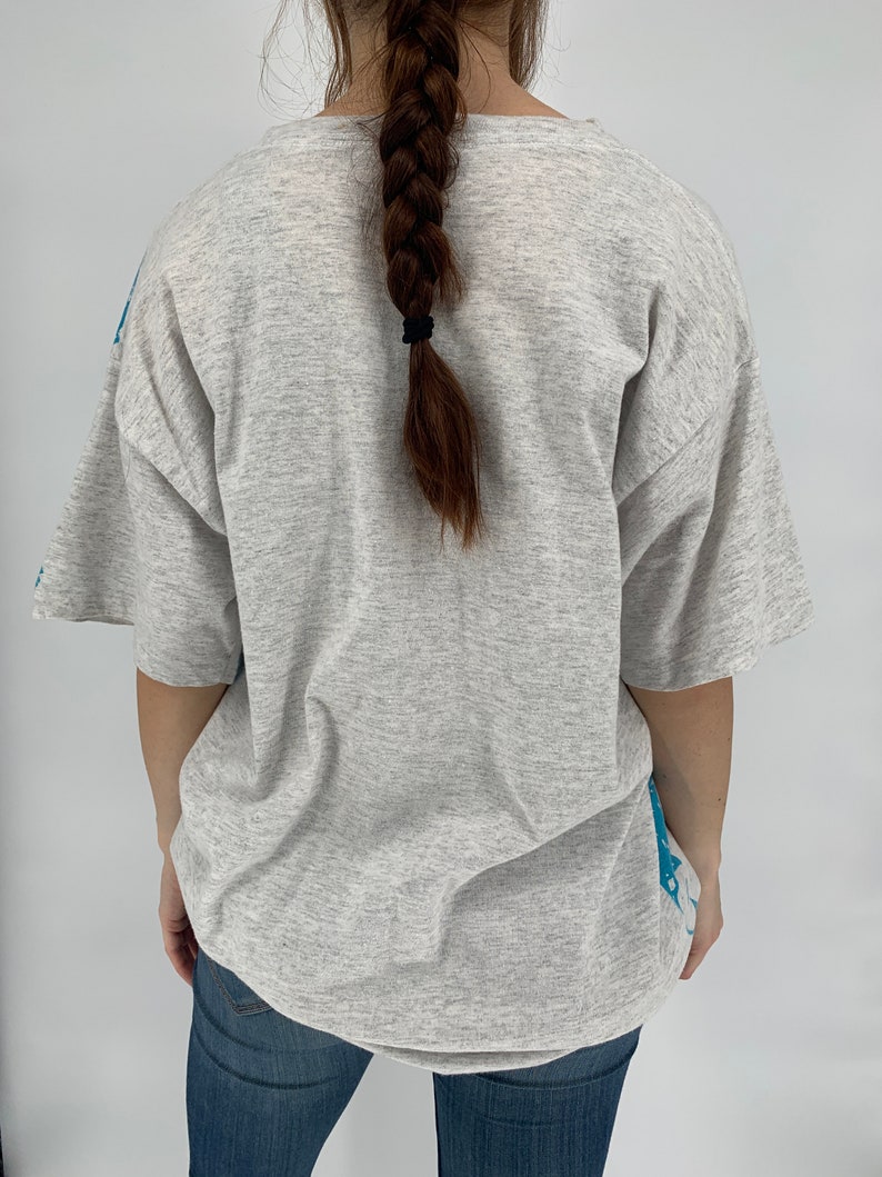 Puede incluir: Una camiseta gris con un estampado gr&aacute;fico azul en la espalda. La camiseta tiene cuello redondo y mangas cortas.