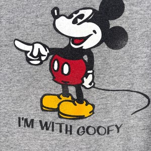 May include: A heather gray t-shirt featuring a screen-printed graphic of Mickey Mouse pointing to the right. Mickey is wearing red shorts and yellow shoes. Below the character, the black text reads I'M WITH GOOFY in a simple font.