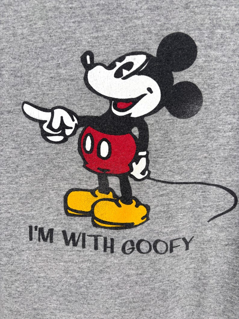 May include: A heather gray t-shirt featuring a screen-printed graphic of Mickey Mouse pointing to the right. Mickey is wearing red shorts and yellow shoes. Below the character, the black text reads I'M WITH GOOFY in a simple font.