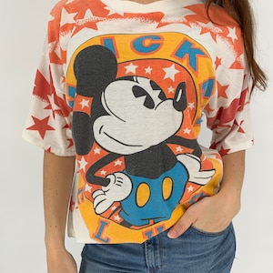 Op de afbeelding: Wit T-shirt met een kleurrijke Mickey Mouse-afbeelding. De afbeelding toont Mickey Mouse in een blauwe en gele outfit met een rode sterrenachtergrond. De tekst "LUCKY" staat in een cirkel rond Mickey Mouse.