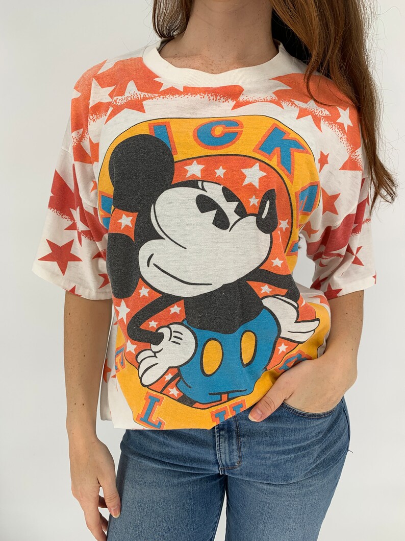 Op de afbeelding: Wit T-shirt met een kleurrijke Mickey Mouse-afbeelding. De afbeelding toont Mickey Mouse in een blauwe en gele outfit met een rode sterrenachtergrond. De tekst "LUCKY" staat in een cirkel rond Mickey Mouse.