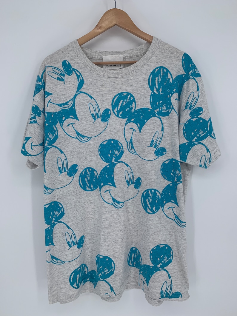 Puede incluir: Una camiseta gris con un estampado azul de Mickey Mouse por todas partes. El estampado de Mickey Mouse es de estilo dibujado.