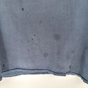 May include: A dark charcoal gray t-shirt featuring a series of small, scattered holes and faint circular stains across the fabric surface. The garment has a visible bottom hem with a double-stitched edge and a plain, solid-colored textile texture.