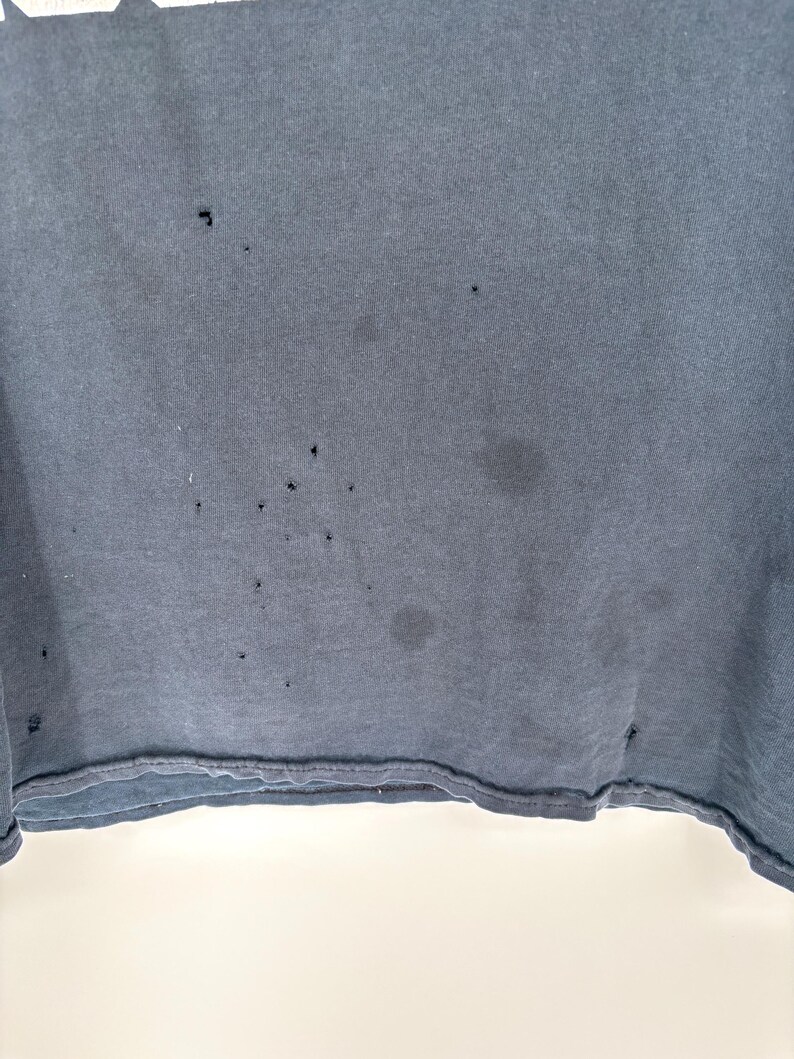 May include: A dark charcoal gray t-shirt featuring a series of small, scattered holes and faint circular stains across the fabric surface. The garment has a visible bottom hem with a double-stitched edge and a plain, solid-colored textile texture.