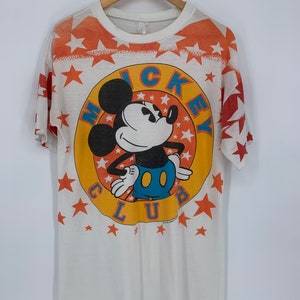 Op de afbeelding: Wit T-shirt met een kleurrijke Mickey Mouse Club-afbeelding. De afbeelding toont Mickey Mouse in een gele cirkel met oranje sterren en de woorden "Mickey Club" eromheen.
