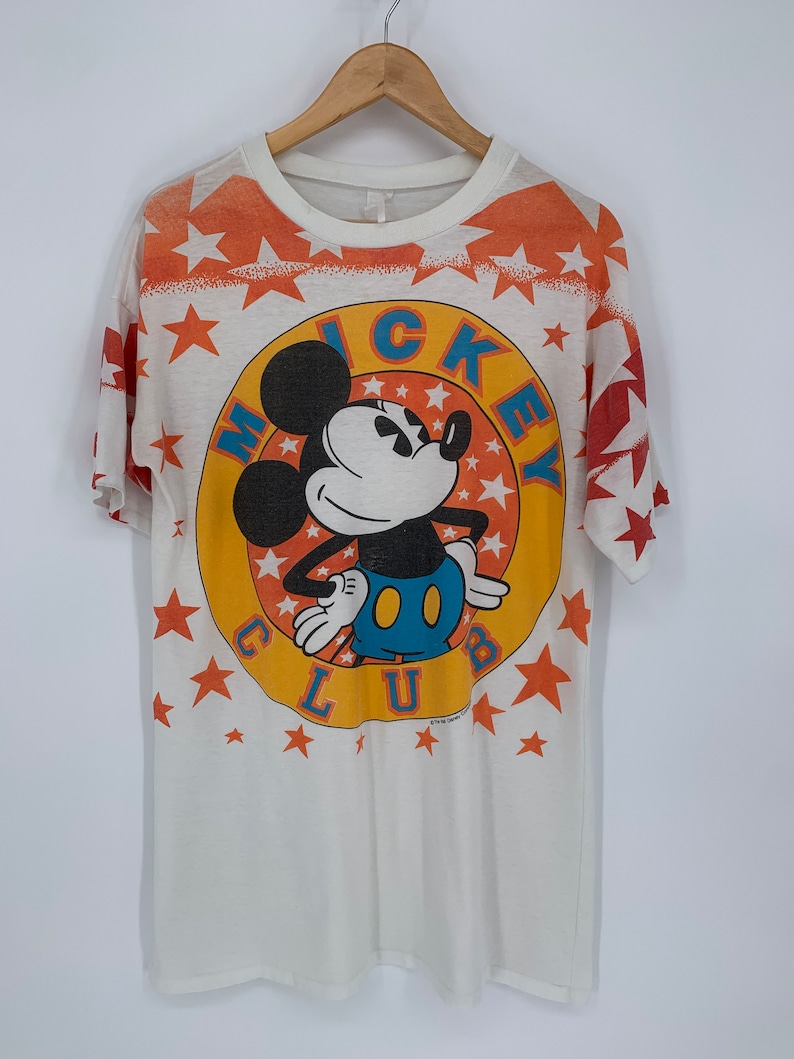 Op de afbeelding: Wit T-shirt met een kleurrijke Mickey Mouse Club-afbeelding. De afbeelding toont Mickey Mouse in een gele cirkel met oranje sterren en de woorden "Mickey Club" eromheen.
