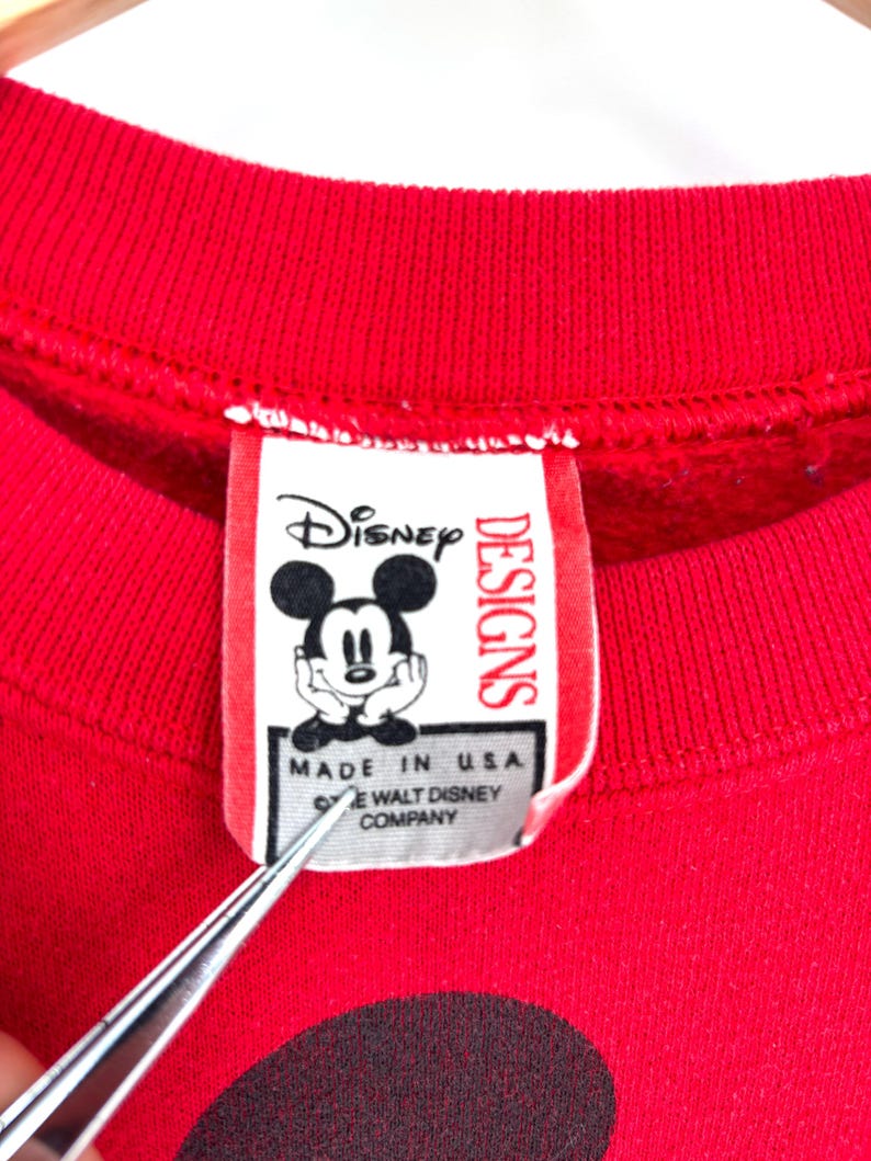 K&ouml;nnte beinhalten: Rotes Sweatshirt mit Mickey Mouse-Grafik. Das Etikett lautet "Disney Designs" und "Made in USA &copy; The Walt Disney Company."