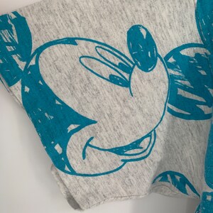 Puede incluir: Camiseta gris con un estampado azul de Mickey Mouse. El estampado es un simple contorno de la cabeza y las orejas de Mickey Mouse.