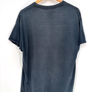 May include: A faded black short-sleeve t-shirt hanging on a wooden hanger. The shirt features a crew neckline, short sleeves with raw-edge hems, and a slightly distressed, worn-in appearance with visible fabric texture and minor signs of wear.