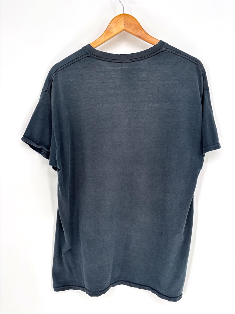 May include: A faded black short-sleeve t-shirt hanging on a wooden hanger. The shirt features a crew neckline, short sleeves with raw-edge hems, and a slightly distressed, worn-in appearance with visible fabric texture and minor signs of wear.